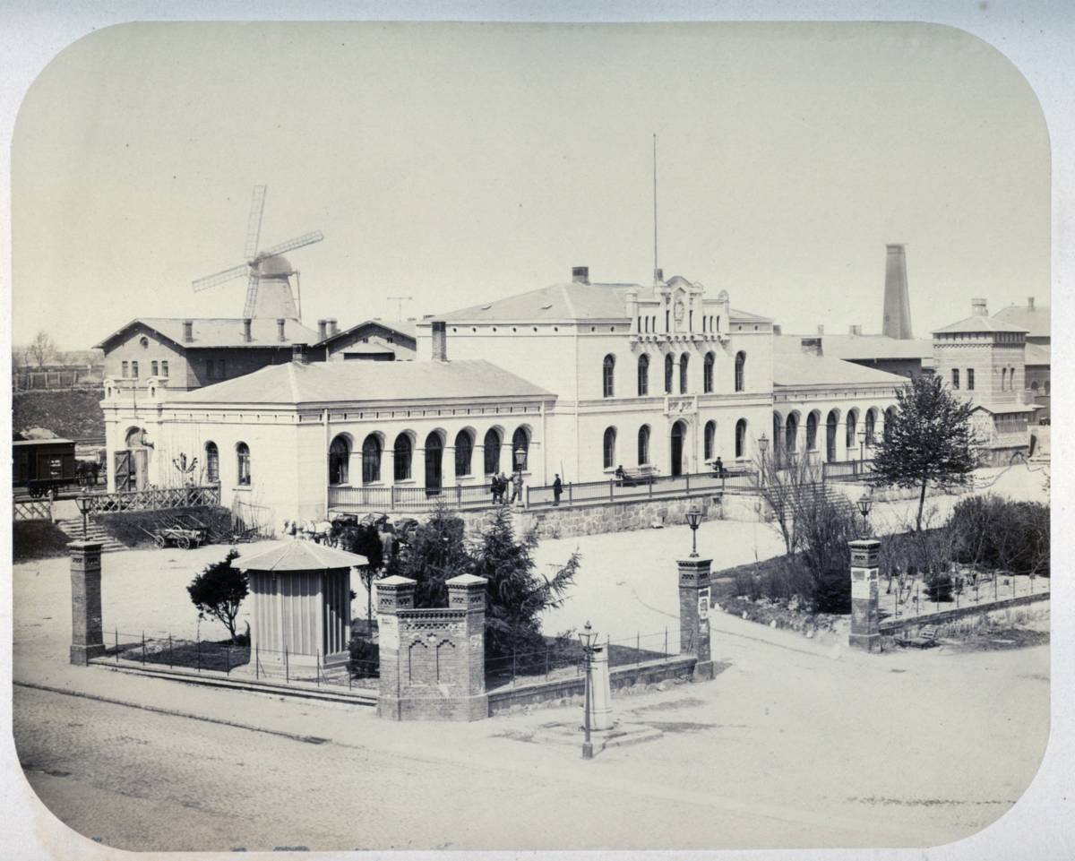 Schwarz-weiß Foto des Schweriner Bahnhofs vor dem Umbau 1888 mit dem Bahnhofsvorplatz, im Hintergrund eine Windmühle. 