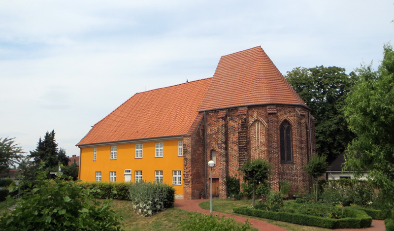 Foto: Chor einer Backsteinkirche, die in ein verputztes gelbes Haus übergeht